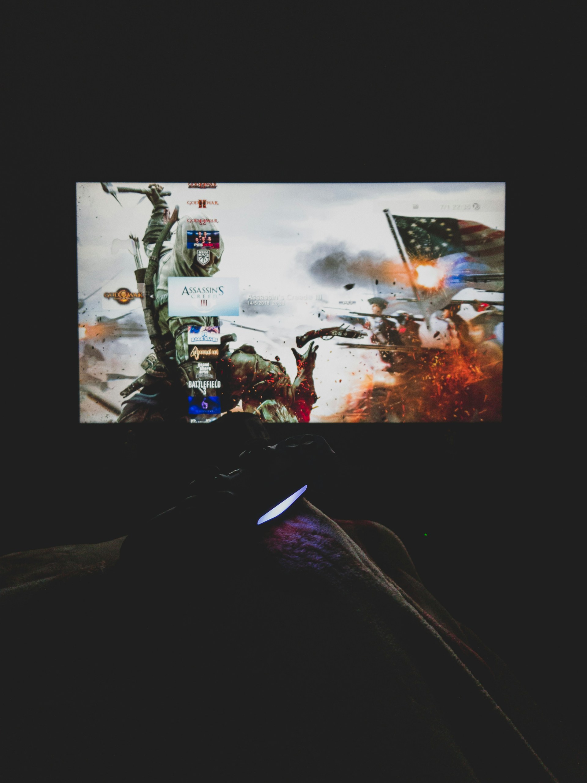 A dimly lit room with a television screen displaying a dynamic, action-packed video game interface featuring a historical battle scene. The foreground shows the silhouette of a gaming console controller held by a person.