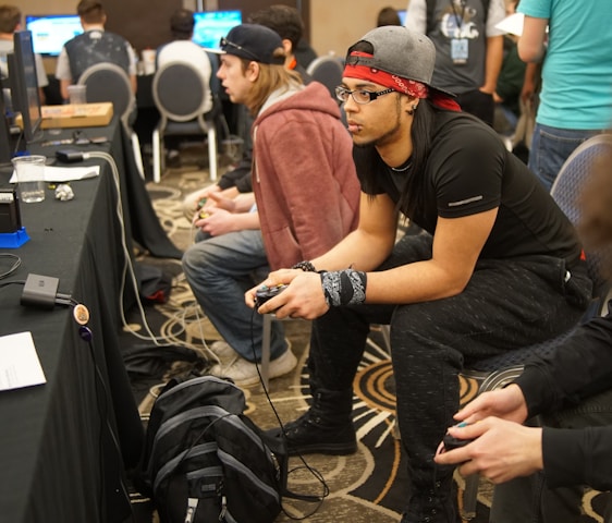Several people are sitting in a room with gaming consoles and monitors, focusing intently on their screens. The setting appears to be a casual gaming event, with participants holding controllers while playing video games. The room has a casual atmosphere with chairs and tables set up for gaming.
