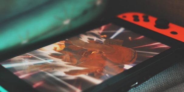 A portable gaming console with neon red and blue controllers displays an action-packed scene from a video game. The screen is focused on a character surrounded by bright, dynamic light effects.