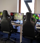 A row of gaming stations equipped with large monitors, gaming chairs, keyboards, and headphones. The setup is organized and ready for gaming, featuring a colorful screen display. The room has a modern and tech-savvy ambiance.
