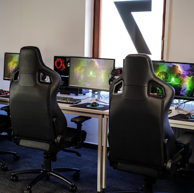 A row of gaming stations equipped with large monitors, gaming chairs, keyboards, and headphones. The setup is organized and ready for gaming, featuring a colorful screen display. The room has a modern and tech-savvy ambiance.