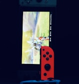 A gaming console with a screen displaying a video game scene, accompanied by two controllers, one colored red and the other blue. The devices are set against a dark backdrop, emphasizing the vibrant colors of the controllers.