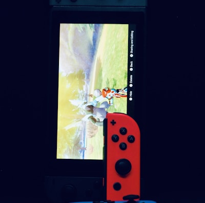 A gaming console with a screen displaying a video game scene, accompanied by two controllers, one colored red and the other blue. The devices are set against a dark backdrop, emphasizing the vibrant colors of the controllers.