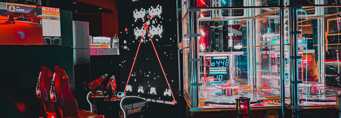 An arcade setting featuring a classic Space Invaders game setup and a modern gaming machine with seats. Bright neon lights illuminate the area, with scores displayed on digital screens. A bright and visually engaging environment full of electronic components.